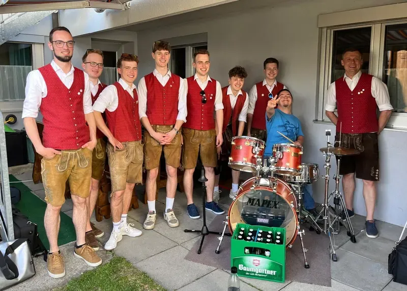 Gruppenfoto der WindBradler in Lederhose und rotem Gilet neben dem Schlagzeug